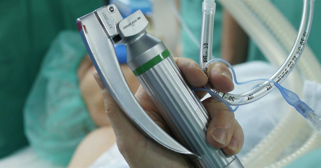 Eine Hand hält medizinische Utensilien. Im Hintergrund liegt ein Patient oder eine Patientin. 